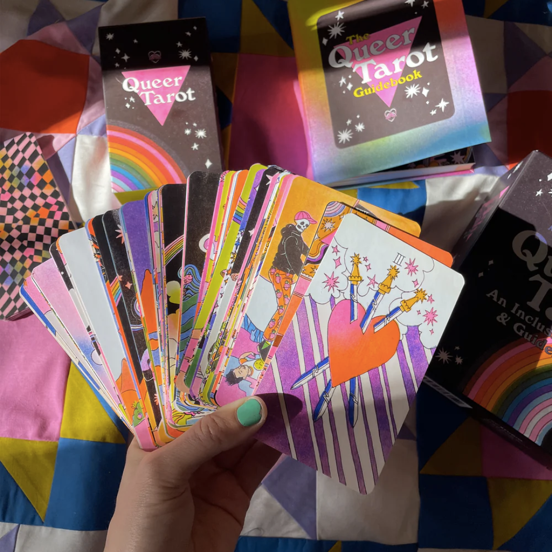 In the foreground, a hand with a painted thumb nail holds the entire deck of cards, which are fanned out to show a small part of each card. In the background, the guidebook and Queer Tarot packaging box are seen. 