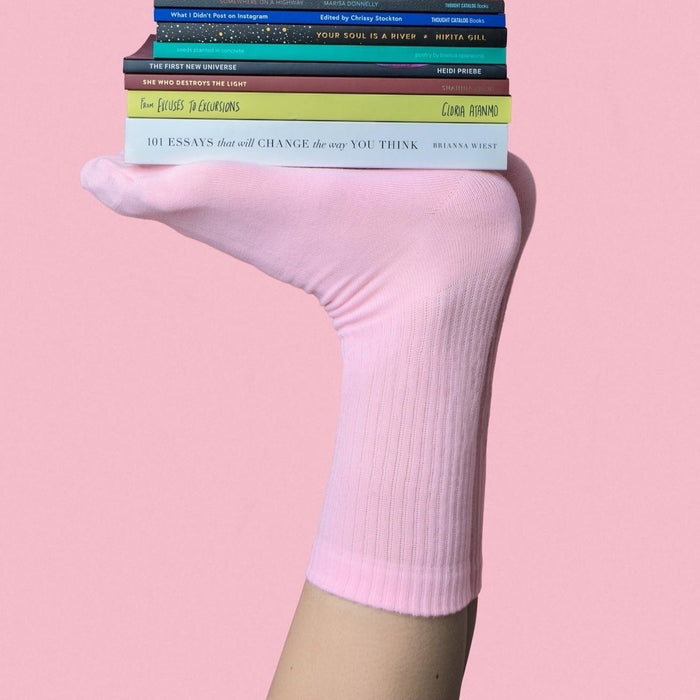 Feet in the air wearing pink socks, balancing a stack of books on top of them. On a pink background