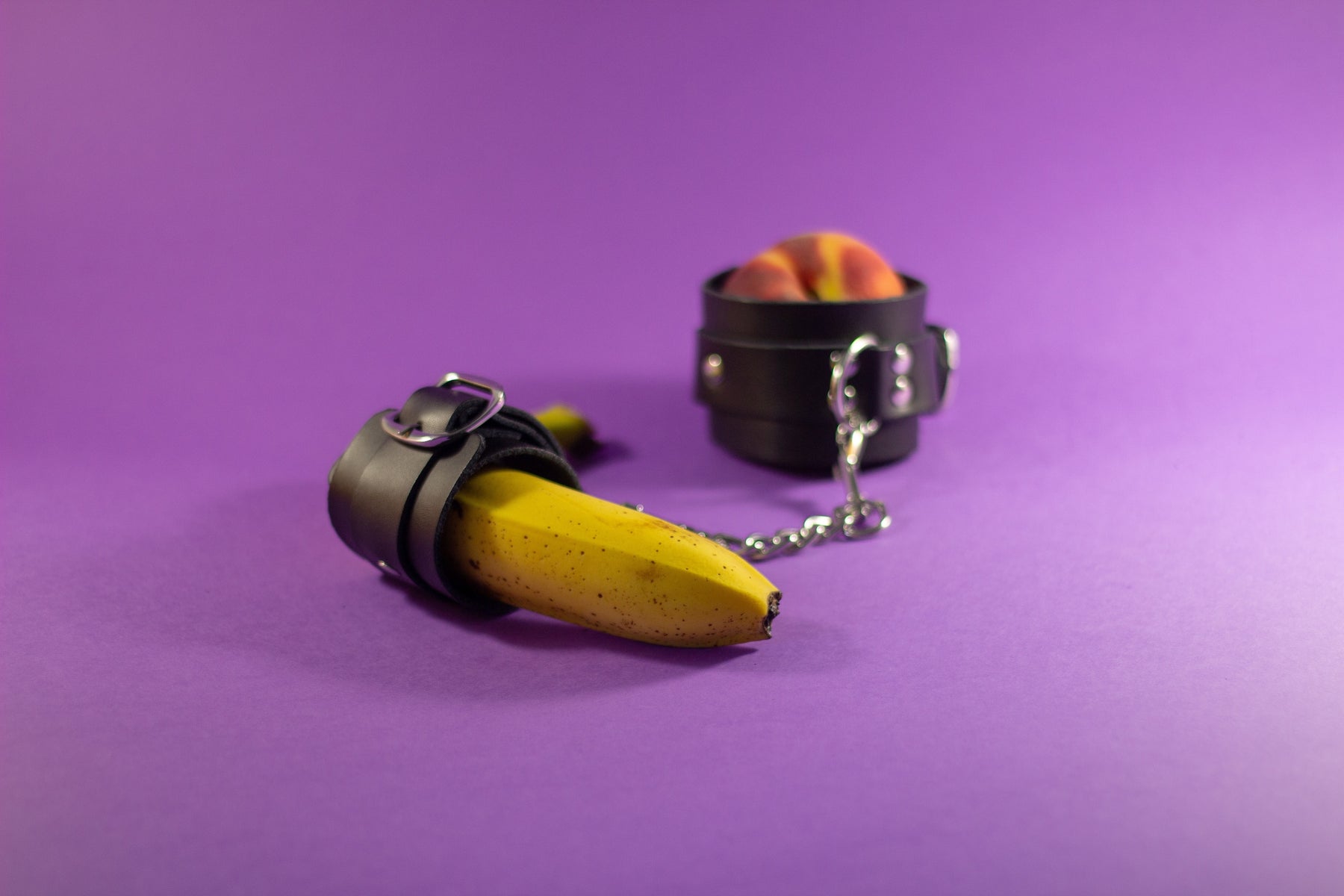 A banana and a peach wearing black restraints on a purple background