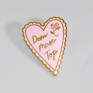 A gold plated enamel pin in the shape of a pink heart that says Dom Masc Top in cursive writing. An etched gold rose is in the top right part of the pin.