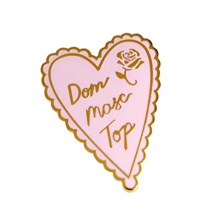 A gold plated enamel pin in the shape of a pink heart that says Dom Masc Top in cursive writing. An etched gold rose is in the top right part of the pin.