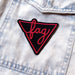 A black triangle patch with neon pink embroidery that says the word "Fag" in cursive. The patch is outlined with a pink triangle, and the triangle conjoins at a cursive F on one side. The patch is seen on a light wash denim jacket pocket.