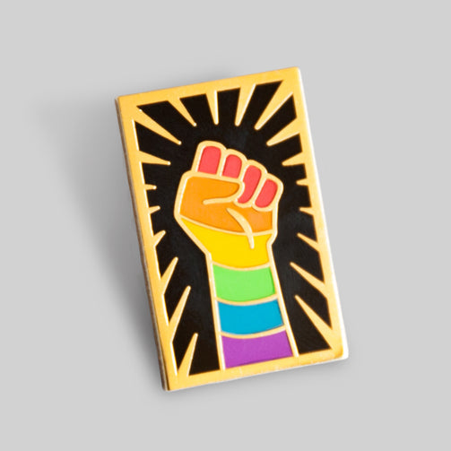 An enamel pin with a forearm and hand making a fist upwards. The arm and hand have the rainbow flag stripes overlayed, and there are gold lines surrounding the fist.