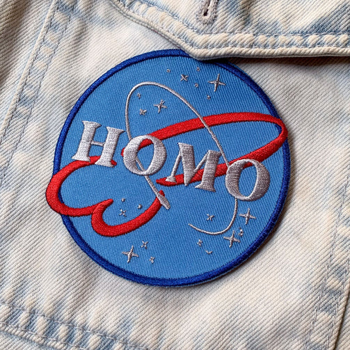 A circular patch that looks like the NASA logo but instead of NASA it says HOMO. The logo has a dark blue outline on a light blue circle. A red swirly design that is shaped like a penis and a finer circular line that looks like the ring of a planet surround the word HOMO. Twinkling stars of various sizes also surround the logo.