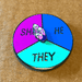 A round pronoun pin, with one third of the circle taken up by the pronoun he, another third of the circle the pronoun she, and the last the pronoun they. A crystal dial that can be spun to different pronouns is in the center of the circle.
