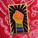 A rectangular patch with a forearm and hand making a fist upwards. The arm and hand have the rainbow flag stripes overlayed, and there are gold lines surrounding the fist. The patch lays on top of a red paisley bandana.