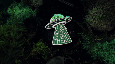 A green spaceship with a beam coming out of it. The words "The Gay is Out There" are written across the beam