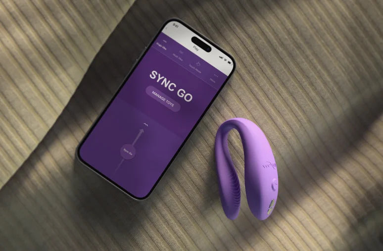 A picture of sync go and a phone with the we-vibe app lying on a bed sheet together 