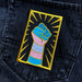 A rectangular patch with a forearm and hand making a fist upwards. The arm and hand have the trans flag stripes overlayed, and there are gold lines surrounding the fist. The patch is seen on a jean jacket pocket, up close. 