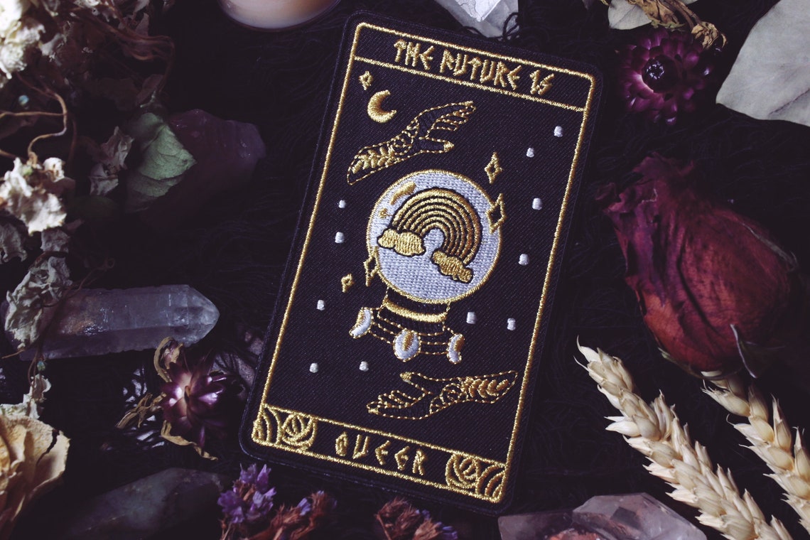 A Tarot card-style patch that is black with gold embroidery. The illustration is a crystal ball with a rainbow in it and sparkles surrounding it. Two hands surround the crystal ball, on the top and bottom. The top of the patch says "The Future is", and the bottom says "Queer".