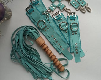 A collection of teal kink equipment sits atop a white table with a vase in the corner with a purple flower in it. The equipment ranges from hand and ankle cuffs with silver buckles, to a flogger with a wooden handle, to small clip attachments with silver clips and buckles. 