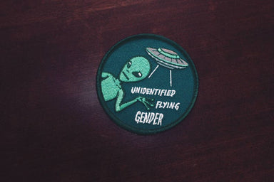 A round teal coloured patch that says "Unidentified Flying Gender" with an illustration of an alien and a flying saucer. 