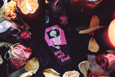A patch shaped like a coffin with a skeleton lying inside on a pillow and pink sheets. The words gay and tired are written at the bottom across the black blanket covering the skeletons lower half.