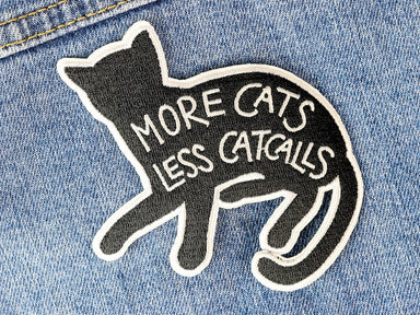 A black patch with white outline shaped like a cat. On the body of the cat are the words "More cats, less catcalls" in white embroidery 