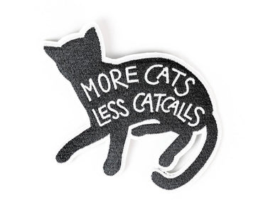 A black patch with white outline shaped like a cat. On the body of the cat are the words "More cats, less catcalls" in white embroidery
