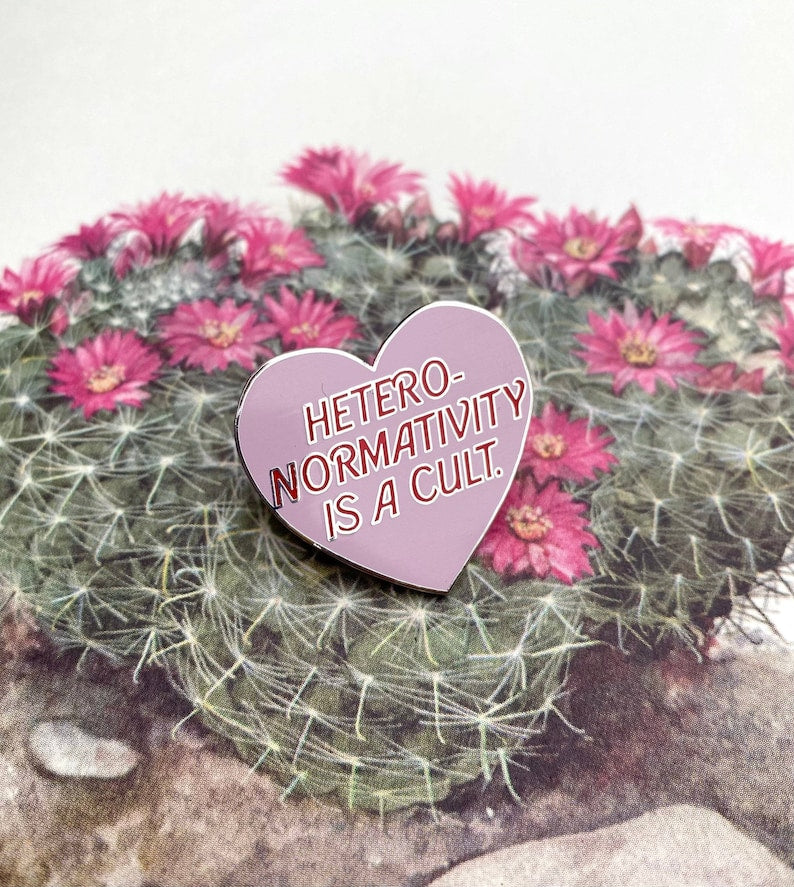 A pink heart shaped enamel pin that says "Heteronormativity is a cult". The pin is displayed on a barrel cactus with pink flowers. 
