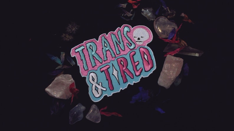 An iron on patch that says Trans & Tired in capital letters. The letter "I" in tired is a four sided star or sparkle and there is a skull closing its eyes next to the word Trans. The patch is coloured with the Trans Flag colours. The patch is surrounded by crystals and flower petals.