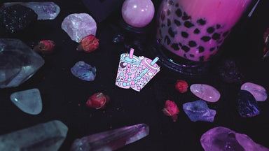 The bubble tea pin is now lying on a table, with crystals and dried rose hip flowers scattered around it. There is a crystal ball and full sized bubble tea in the background of the photo.