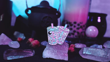 An enamel pin depicting two pink, blue, and white bubble teas. One bubble tea has a carving of Saturn in it and the other has a carving of a crescent moon. The pin is balancing on a crystal, and in the background of the picture there are more crystals, a crystal ball, a large cup of bubble tea with boba bubbles, and a cauldron with a pentacle on it. 