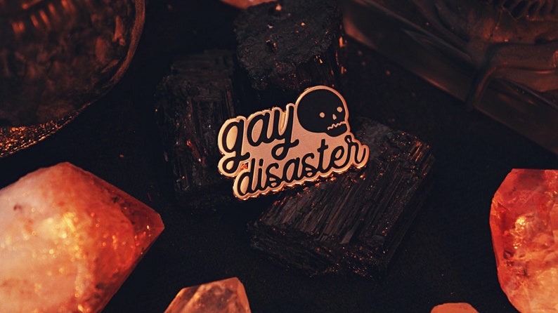 A gold plated enamel pin with black writing that says "Gay Disaster" in lowercase, half-cursive writing. Next to the word gay is a simple illustration of a skull with a straight faced expression. The pin is surrounded with crystals. 