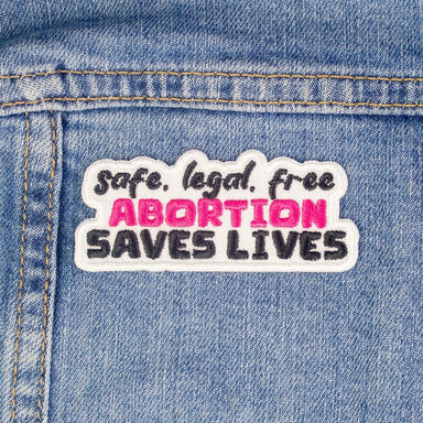 A patch with a white background and black text that reads "safe, legal, free abortion saves lives". The word abortion is in pink