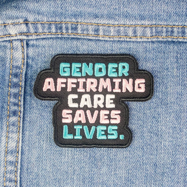 A patch with a black background and text that says "Gender affirming care saves lives". The text is the colours of the transgender flag. 