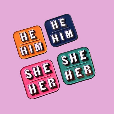 Four square enamel pins with raised block letters, arranged on a pink background. The top-left pin is orange and reads 'HE HIM', the top-right is navy and reads 'HE HIM', the bottom-left is pink and reads 'SHE HER', and the bottom-right is teal and reads 'SHE HER'.