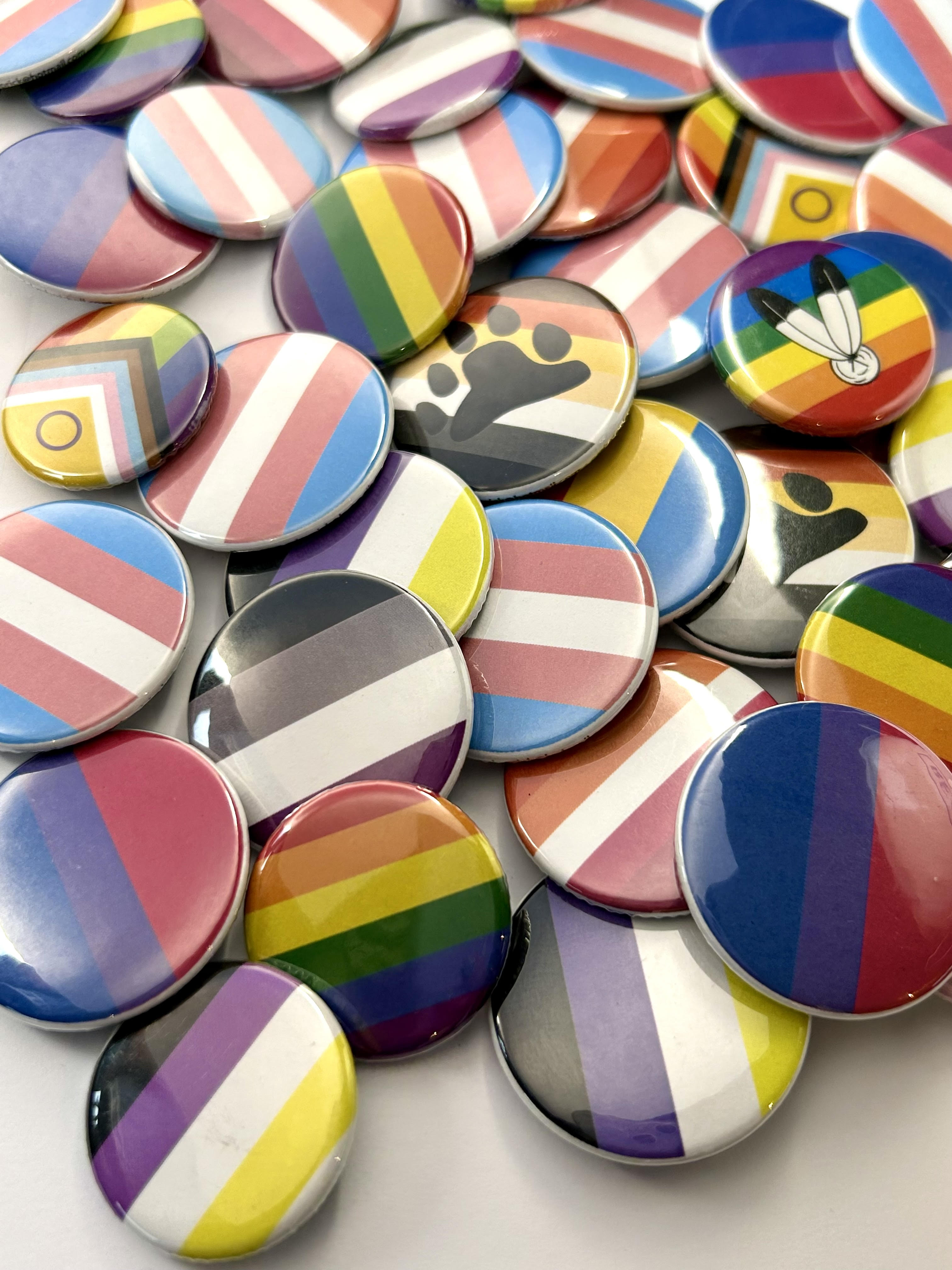 An assortment of round buttons with different Pride flags on them are scattered across a white background.