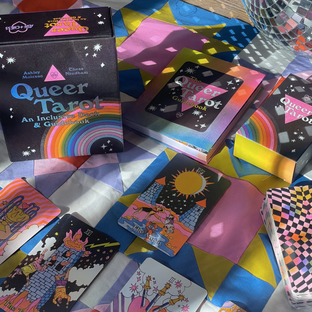 Various tarot cards, a guide book that says "Queer Tarot" and the packaging of the tarot deck set lay across a colourful blanket. The cards are illustrated in bold colours of pink, yellow, black, and orange. A disco ball peeks into the top right corner and casts reflective light across the cards and blanket. 