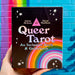 A black box with the text "Queer Tarot: An Inclusive Deck and Guidebook" is held up to a painted brick wall. There is a rainbow with all the progress pride flag colours in the bottom right corner on the box, and twinkly star illustrations over the cover. The brick wall in the background is blue with a rainbow painted on.